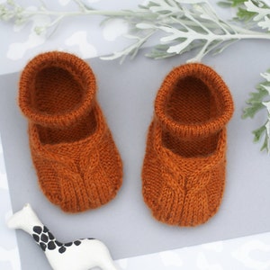 May include: A pair of knitted baby booties in a burnt orange colour. The booties have a cable knit pattern and a fold-over cuff.