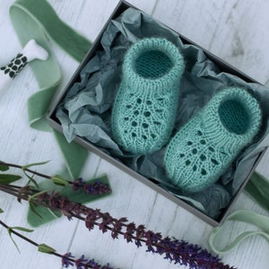 May include: A pair of teal blue knitted baby booties with a lacy pattern, displayed in a gift box with tissue paper.
