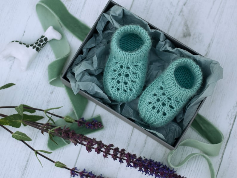 May include: A pair of teal blue knitted baby booties with a lacy pattern, displayed in a gift box with tissue paper.