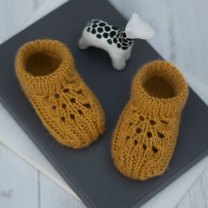 May include: A pair of knitted baby booties in a mustard yellow color with a lacy pattern. The booties are sitting on a gray surface.