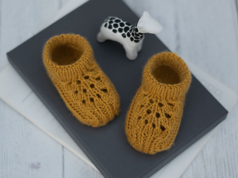May include: A pair of knitted baby booties in a mustard yellow color with a lacy pattern. The booties are sitting on a gray surface.