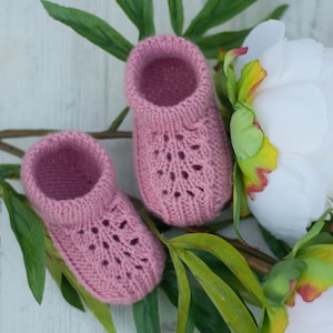 May include: A pair of pink knitted baby booties with a lacy design, resting on a white surface with green leaves and white flowers.