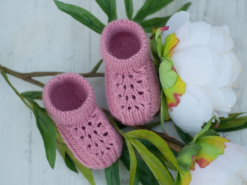 May include: A pair of pink knitted baby booties with a lacy design, resting on a white surface with green leaves and white flowers.