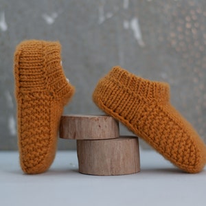 Könnte beinhalten: Zwei Paar gestrickte Baby-Schühchen in Senfgelb. Die Schühchen sind mit einem strukturierten Strickmuster gefertigt und haben einen umgeschlagenen Bund.