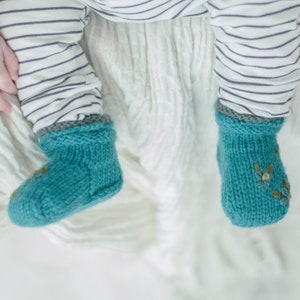 May include: A pair of teal blue knitted baby booties with a floral design on one bootie. The booties are on a white background.