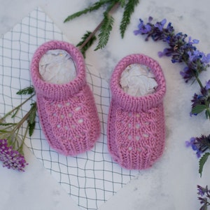 May include: A pair of pink knitted baby booties with a lacy design. The booties are sitting on a white surface with a grid pattern.