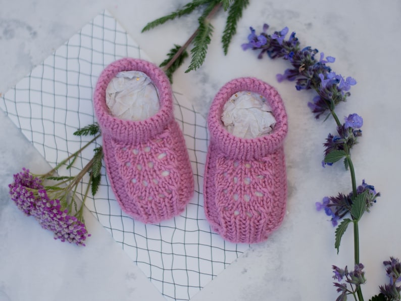 May include: A pair of pink knitted baby booties with a lacy design. The booties are sitting on a white surface with a grid pattern.