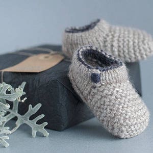 Könnte beinhalten: Ein Paar graue gestrickte Babyschuhe mit dunkelblauem Besatz. Die Schuhe liegen auf einem schwarzen Geschenkbox mit einem braunen Etikett.