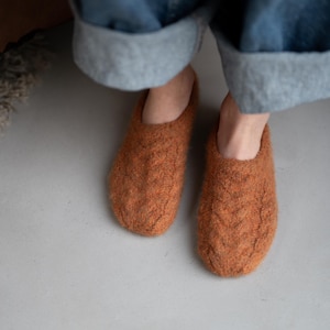 May include: A pair of burnt orange slippers featuring a textured cable knit pattern. The slippers have a rounded toe design and a slip-on style, crafted from a fuzzy, wool-like material that covers the top and sides of the footwear.