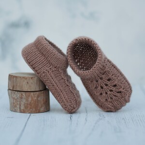 May include: A pair of knitted baby booties in a light brown color. The booties have a delicate lace pattern and are made with soft yarn.