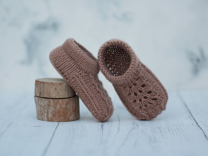 May include: A pair of knitted baby booties in a light brown color. The booties have a delicate lace pattern and are made with soft yarn.