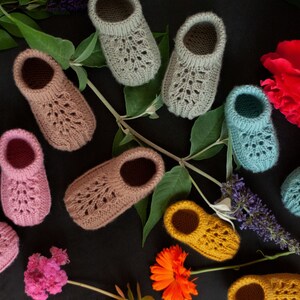 May include: A collection of ten knitted baby booties in various colors, including pink, blue, green, and yellow. The booties are laid out on a black background with flowers and greenery.