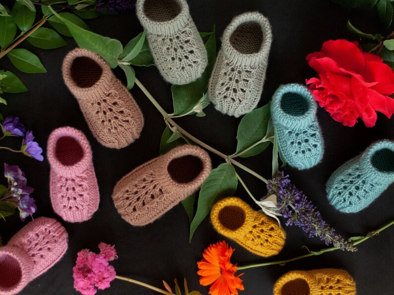 May include: A collection of ten knitted baby booties in various colors, including pink, blue, green, and yellow. The booties are laid out on a black background with flowers and greenery.