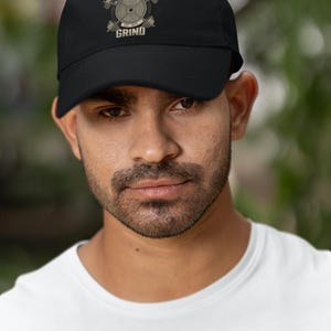 Puede incluir: Gorra de béisbol negra con el texto "EMBRACE THE GRIND" y un gráfico de levantamiento de pesas. La gorra está hecha de un material duradero y presenta una visera curva. El diseño incluye las palabras "Old School Iron Club" y el año "EST. 1974".