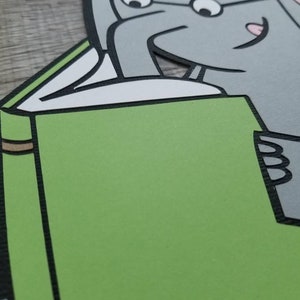 May include: A layered paper cutout featuring a gray elephant character with glasses reading a large green book. A small pink pig character is visible at the top of the elephant's head. The design uses bold black outlines and solid colors.