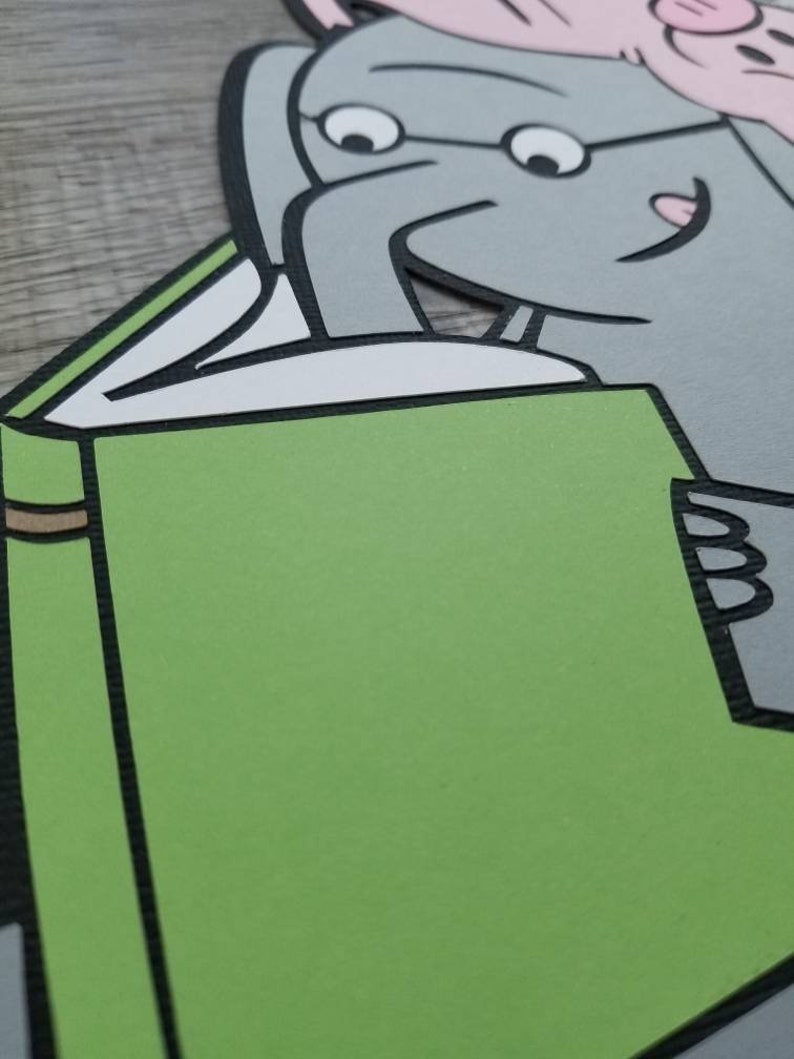 May include: A layered paper cutout featuring a gray elephant character with glasses reading a large green book. A small pink pig character is visible at the top of the elephant's head. The design uses bold black outlines and solid colors.