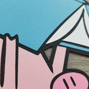 May include: A layered paper cutout featuring a stylized pink pig figure with a black outline, positioned beneath a light blue and white geometric shape. The design uses flat, solid colors with a distinct black border separating each paper element.