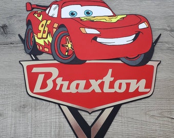 May include: Red cartoon race car with a number 95 on the side and a smiling face. The car is on top of a red and gold shield with the name Braxton written in gold letters.