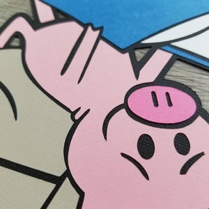 May include: A layered paper cutout featuring a pink pig with a dark pink snout, positioned above a beige shape and holding a blue rectangular object with a white corner. The design uses bold black outlines to define the pig and the geometric shapes.