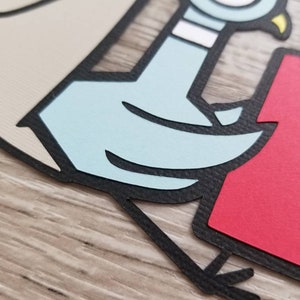 May include: A layered paper craft cutout featuring a light blue cartoon pigeon with a white neck band and a yellow beak. The pigeon is outlined in black and positioned next to a solid red rectangular shape on a light beige background.