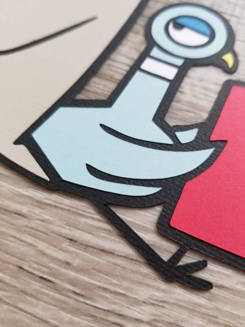 May include: A layered paper craft cutout featuring a light blue cartoon pigeon with a white neck band and a yellow beak. The pigeon is outlined in black and positioned next to a solid red rectangular shape on a light beige background.