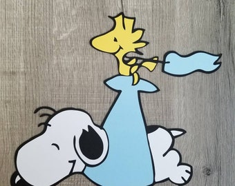 May include: A layered paper cutout featuring a white dog with black ears and a black nose lying down, wearing a light blue blanket. A small yellow bird stands on the blanket, holding a light blue flag. The design has bold black outlines.