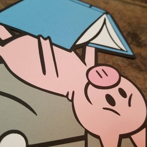 May include: A layered paper cutout featuring a pink pig character with black outlines, positioned above a gray shape. The pig is holding a light blue book with white pages. The design uses flat colors and bold black lines to define the shapes.