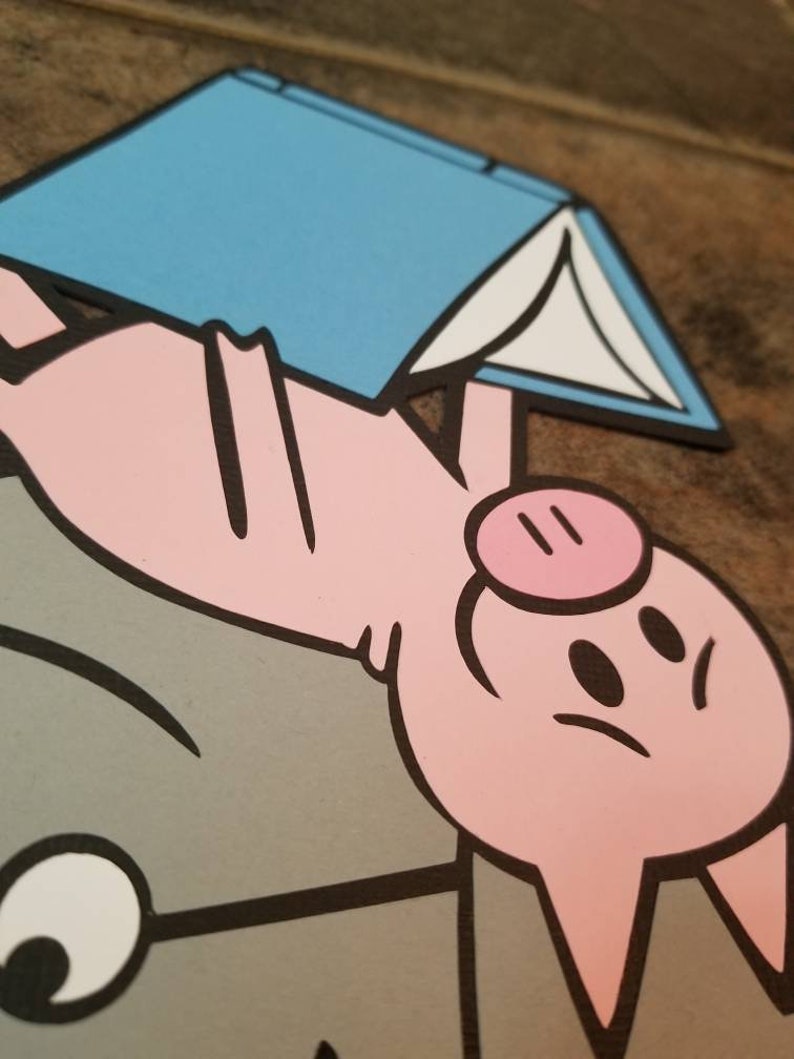 May include: A layered paper cutout featuring a pink pig character with black outlines, positioned above a gray shape. The pig is holding a light blue book with white pages. The design uses flat colors and bold black lines to define the shapes.
