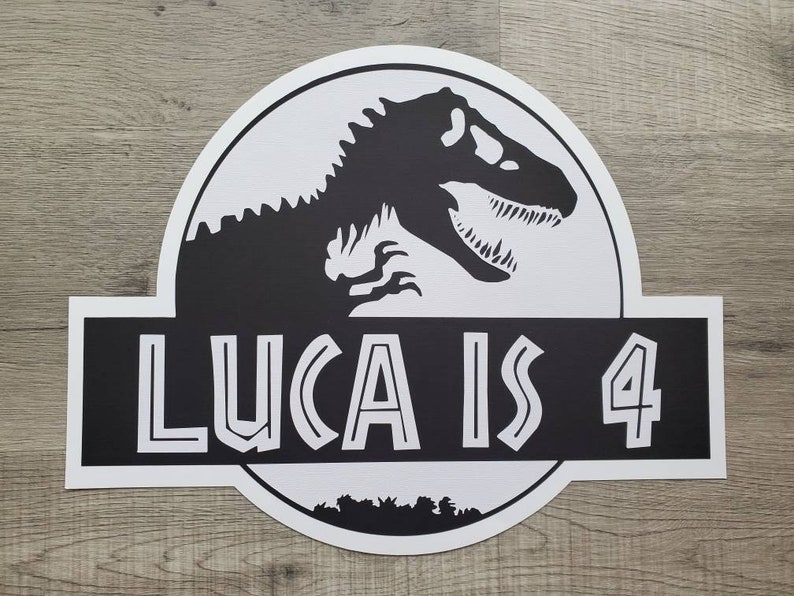 May include: Black and white Jurassic Park-themed birthday sign with a dinosaur silhouette and the text "Luca is 4".