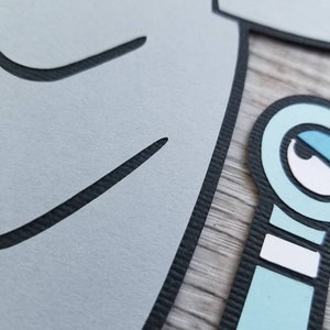 May include: A layered paper cutout featuring a large light gray shape with black outlines and a smaller light blue character element with a white eye, black pupil, and black outlines, showcasing a textured surface with visible paper edges.
