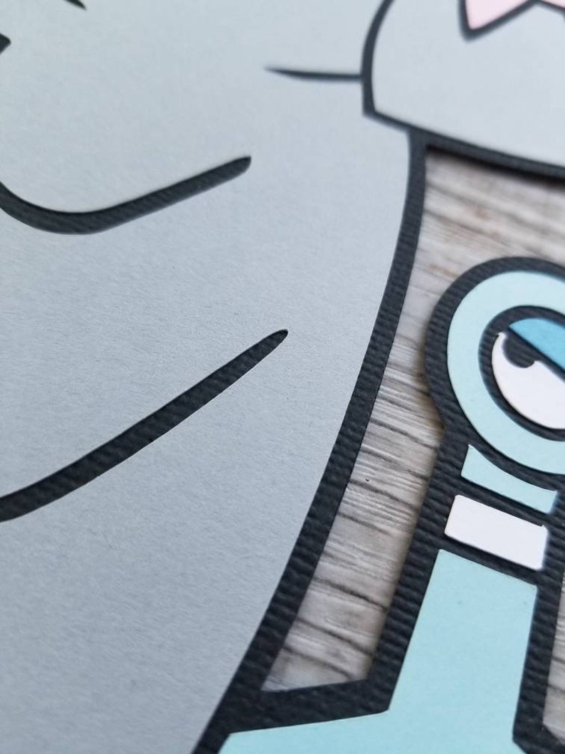 May include: A layered paper cutout featuring a large light gray shape with black outlines and a smaller light blue character element with a white eye, black pupil, and black outlines, showcasing a textured surface with visible paper edges.