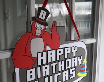 May include: A red and gray birthday sign with the text "HAPPY BIRTHDAY LUCAS". The sign features a cartoon monkey wearing a top hat and a party hat. A red ribbon is attached for hanging.
