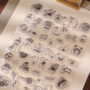 Könnte beinhalten: Klarer Stempel-Set mit Illustrationen zu Outdoor- und Freizeitaktivitäten, darunter ein Segelboot, ein Zelt und ein Grill. Eine goldfarbene, buchförmige Büroklammer und eine dekorative Feder sind ebenfalls vorhanden.