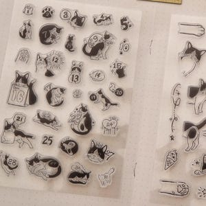 以下が含まれることがあります： 様々な猫のイラストと数字が描かれたクリアスタンプセット。様々なポーズの猫、リボン、足跡、そして「Important」、「Remember」、「Check！」の文字が含まれています。ジャーナリングやクラフトに最適です。