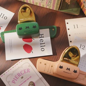 Op de afbeelding: Een verzameling briefpapierartikelen, waaronder een groene en perzikkleurige perforator, met gouden accenten en kattenillustraties. De afbeelding toont ook kaarten met het woord "Hello" en kalenderpagina's. Dozen met de tekst "PIERCER" en "HOLE PUNCH" zijn ook zichtbaar.