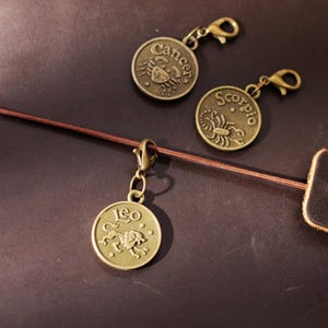 May include: Bronze-colored zodiac charms with lobster clasps, featuring the Cancer, Scorpio, and Leo symbols. The charms are displayed on a brown leather journal with a matching closure.