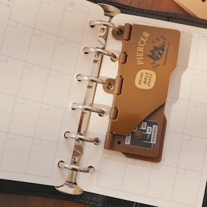 Könnte beinhalten: Offener schwarzer Lederplaner mit monatlicher Kalenderansicht. Ein brauner "PIERCER" Mini-Locher ist an den Metallringen des Planers befestigt. Zusätzliche grüne und beige Locher sind sichtbar.