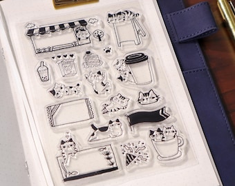 May include: Clear stamp set featuring various cat-themed illustrations, including cats, food, and decorative elements. The stamps are displayed on a white background, held within a blue notebook. A gold-colored clip and pen are also visible.