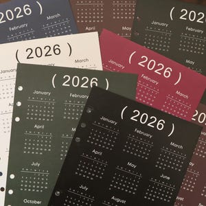Peut inclure: Plusieurs pages de calendrier 2026 de différentes couleurs, dont noir, bordeaux, vert foncé et blanc. Chaque page affiche les mois de l'année, avec l'année 2026 en évidence. Les pages sont perforées pour la reliure.