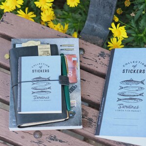 May include: A collection of notebooks and stickers with a Lisbon Fish Market theme. The notebooks are black and light blue, with the text "COLLECTION OF STICKERS" and "Sardines" printed on them. A green pen is attached to the notebook.