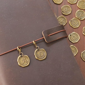 May include: A brown leather journal with a leather strap closure and two bronze letter charms. Additional bronze letter charms are scattered on a wooden surface. The charms are in the shape of a wax seal.