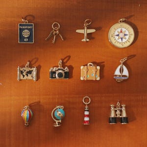 May include: A collection of twelve gold-toned metal travel-themed charms including a blue passport, keys, an airplane, a compass, a map, a camera, a suitcase, a sailboat, a hot air balloon, a globe, a lighthouse, and a pair of binoculars.