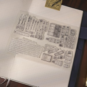 Puede incluir: Una hoja de sellos de silicona transparente con ilustraciones de línea negra de diversos materiales de arte, incluyendo pinceles, espátulas, tubos de pintura, una regla y una escuadra. Se incluye un bloque de texto sobre Joseph Mallord William Turner.