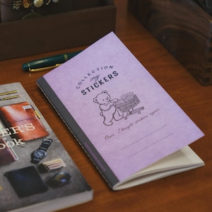 以下が含まれることがあります： 「COLLECTION my STICKERS」の文字と、買い物カートを押すクマのイラストが描かれた紫色のノート。 ノートは開かれており、白いページが見える。 「TRAVELER'S notebook」と書かれた別のノートも見える。