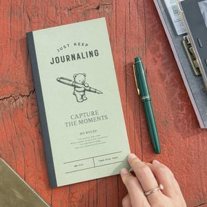 May include: A light green journal with a black spine featuring a bear illustration holding a pen, the text "JUST KEEP JOURNALING" and "CAPTURE THE MOMENTS", and a dark green fountain pen resting beside it on a textured reddish-brown surface.
