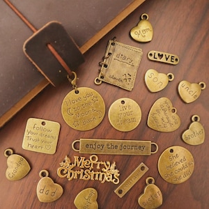 Puede incluir: Una colección de colgantes antiguos de bronce con varias formas e inscripciones. Los colgantes incluyen corazones, círculos y rectángulos, con frases como "I love you to the Moon & Back" y "Merry Christmas". Están dispersos sobre una superficie de madera.