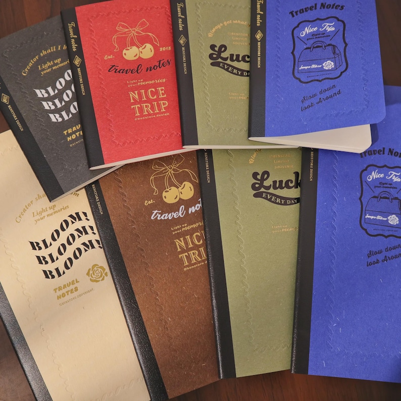 May include: A collection of travel notebooks in various colors, including red, brown, green, and blue. Each notebook features embossed designs and text such as "Travel Notes," "Nice Trip," and "Luck Every Day." The notebooks are arranged on a wooden surface.