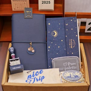 Op de afbeelding: Een verzameling briefpapier in een houten doos. Bevat een marineblauw notitieboek met een fietsbedel, een portemonnee met een maan- en sterrenpatroon en een stempel met de tekst "PAR AVION". Bevat ook een stempelkussen en een papier met "nice trip".