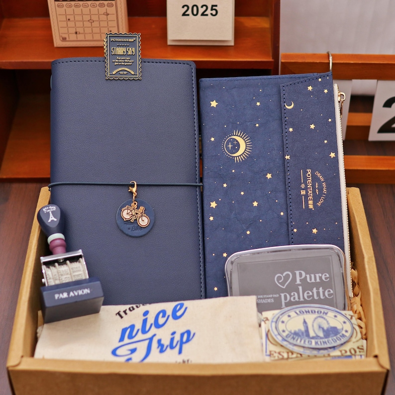 Op de afbeelding: Een verzameling briefpapier in een houten doos. Bevat een marineblauw notitieboek met een fietsbedel, een portemonnee met een maan- en sterrenpatroon en een stempel met de tekst "PAR AVION". Bevat ook een stempelkussen en een papier met "nice trip".