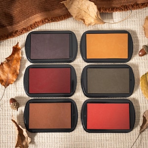 Könnte beinhalten: Sechs rechteckige Stempelkissen in schwarzen Gehäusen, jedes gefüllt mit einem anderen Farbton von Herbstfarben: Braun, Orange, Rot, Olivgrün und Rost. Die Kissen sind auf einer beigen Oberfläche mit Herbstblättern und Eicheln angeordnet.